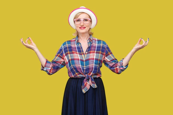 calm modern stylish mature woman in casual style with hat and eyeglasses standing with raised arms while doing yoga pose exercises on yellow background - Photo, Image