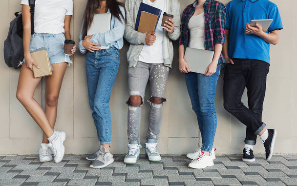 Adolescentes em pé na parede do campus, segurando dispositivos e livros
 - Foto, Imagem