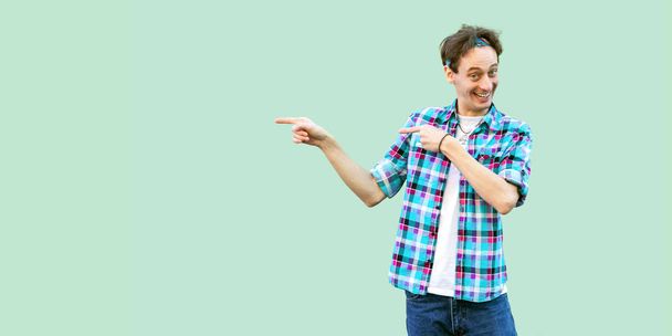 happy funny young man in casual blue checkered shirt and headband standing with satisfied face and showing at wall on green background - Φωτογραφία, εικόνα