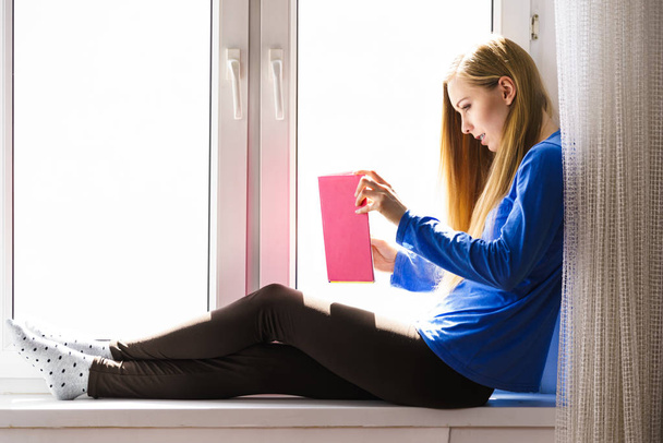 Woman sitting on window sill reading book at home - Foto, Imagen