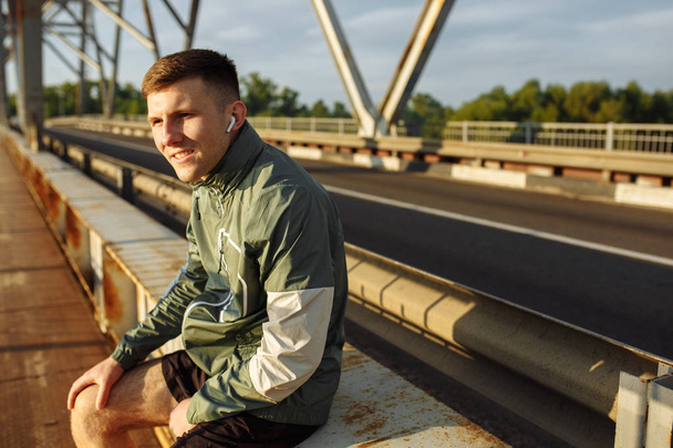 Jeune homme en tenue de sport assis sur un pont et écoutant de la musique dans les écouteurs
 - Photo, image