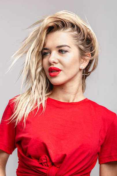 Young blond girl with a red  lipstick short  in a red t-shirt poses staying against a wall in the studio  - Fotó, kép
