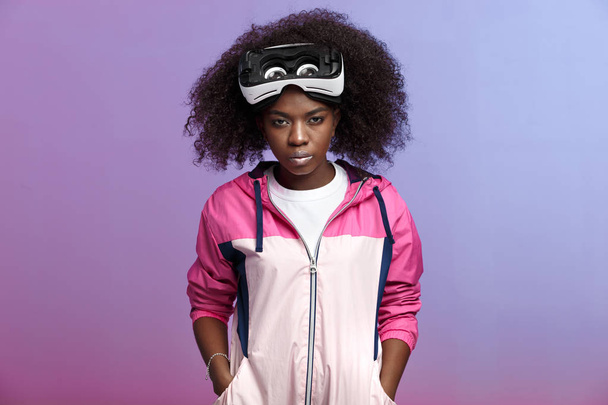 Mod curly brown-haired girl dressed in the pink sports jacket wearing on her head the virtual reality glasses poses in the studio on neon background - Zdjęcie, obraz