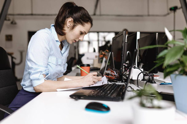 Jonge vrouw gekleed in kantoor stijl kleding werkt op de laptop en het maken van notities in het document zitten aan het bureau in het moderne kantoor - Foto, afbeelding