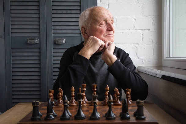 Ένας γέρος κάθεται μπροστά σε μια σκακιέρα, αναρωτιέται και κοιτάζει έξω από το παράθυρο πριν το παιχνίδι - Φωτογραφία, εικόνα