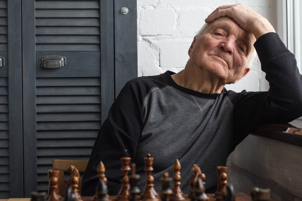 Ένας γέρος κάθεται μπροστά σε μια σκακιέρα, σκέφτεται και κοιτάζει ευθεία μπροστά - Φωτογραφία, εικόνα