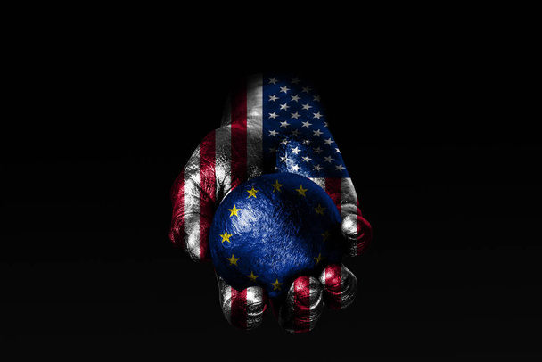 Una mano con una bandera de EE.UU. dibujada sostiene una pelota con una bandera de la UE dibujada, un signo de influencia, presión o conservación y protección
. - Foto, imagen