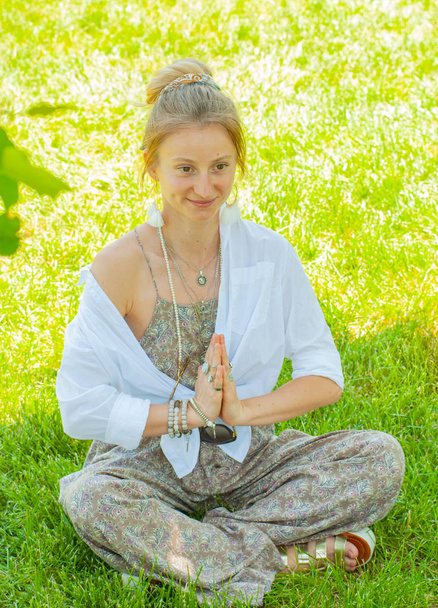Happy woman is meditating sitting in Lotus pose on grass. Beautiful boho style woman with accessories enjoying sunny day in park. - Photo, Image