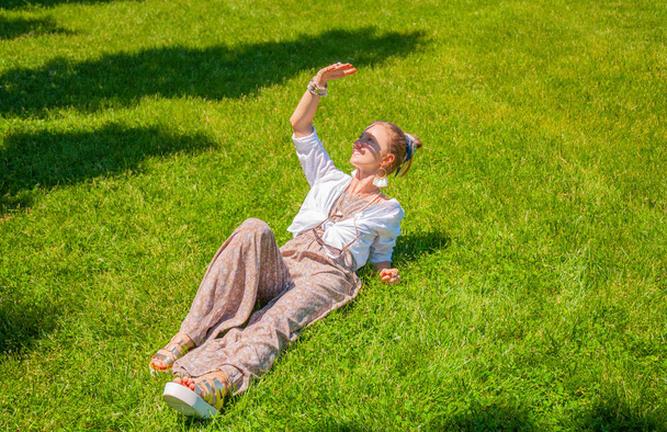 Beautiful boho style woman with accessories is enjoying summer sunny day on grass in park. - Photo, Image