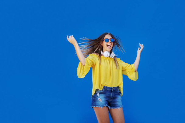 Joven adolescente caucásica alegre en blusa amarilla y pantalones cortos de mezclilla saltando frente al fondo azul
. - Foto, Imagen