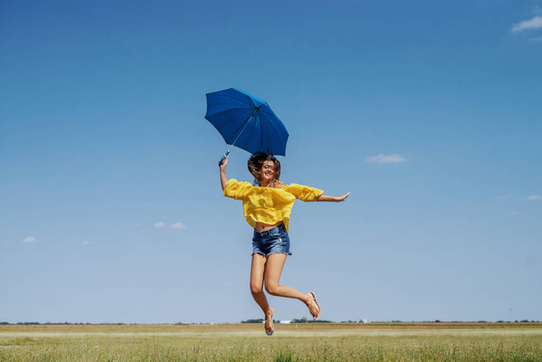Cheerful Caucasian teenage girl in yellow blouse, denim shorts and with eyeglasses jumping outdoor with blue umbrella in hand. - Photo, Image