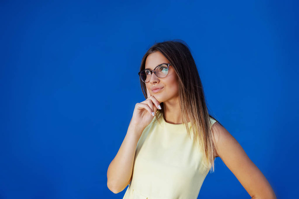 Portrait of beautiful pensive Caucasian teenage girl in yellow dress standing in front of blue background. - Photo, Image