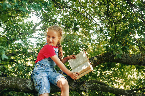 Klein blondine meisje met een boek op een boom. Schoolonderwijs voor kinderen - Foto, afbeelding
