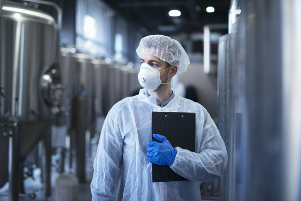 Technoloog in beschermend wit pak met smoothing en masker staande in voedsel fabriek. - Foto, afbeelding
