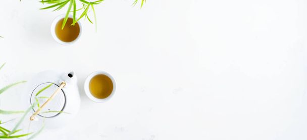 Two white cups of tea and teapot surrounded with green leaves on white background - Foto, afbeelding