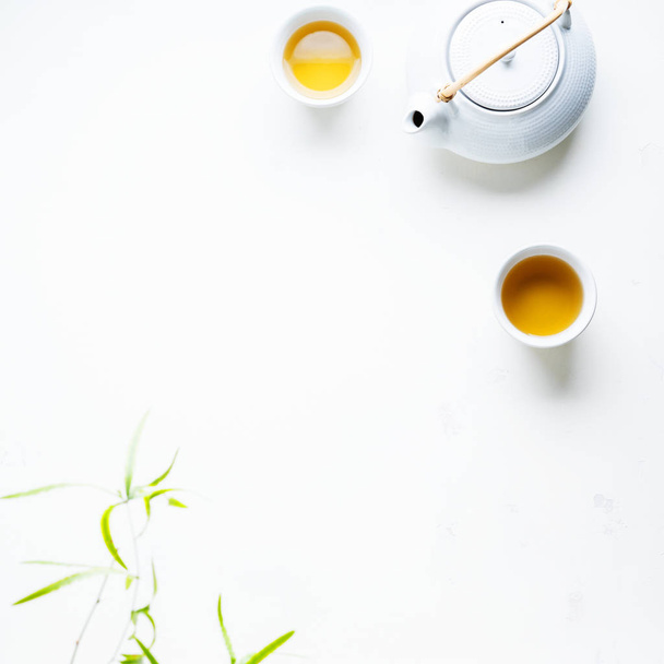 Two white cups of tea and teapot surrounded with green leaves on white background - Фото, изображение