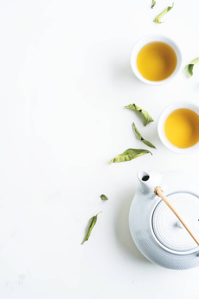 Two white cups of tea and teapot surrounded with green leaves on white background - Фото, зображення