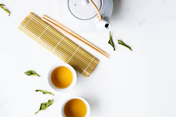 White cups of tea, bamboo mat and teapot surrounded with green tea dry leaves on white background - Fotografie, Obrázek