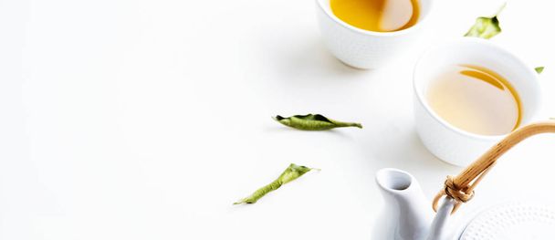 Two white cups of tea and teapot surrounded with green leaves on white background - Foto, Imagen