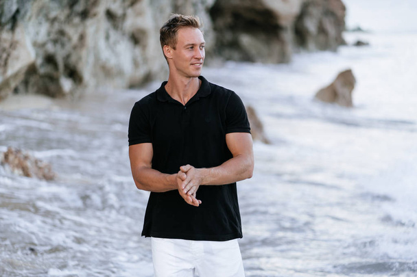 Sexy smiling man in black t-shirt resting on beach, ocean waves at background.  - Foto, Imagem