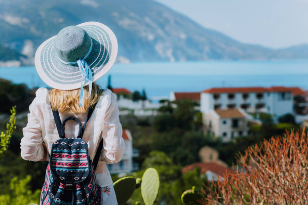 Vintage photo of tourist female with blue sun hat and travel backpack enjoying panorama of colorful small Assos village. Kefalonia, Greece - Foto, imagen
