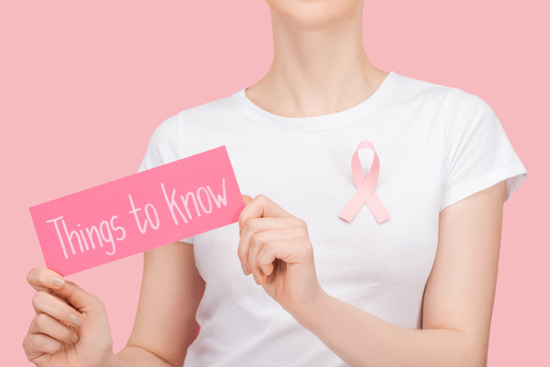 περικομμένη όψη της γυναίκας με ροζ σύμβολο του καρκίνου του μαστού κρατώντας ροζ κάρτα με πράγματα για να γνωρίζουν γράμματα απομονώνονται σε ροζ - Φωτογραφία, εικόνα