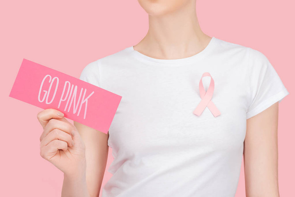 oříznutý pohled na ženu s růžovým náznakem rakoviny prsu s růžovou kartou s růžovým písmem v izolovaném růžovém - Fotografie, Obrázek