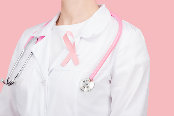 částečný pohled na lékaře v bílém kabátě s růžovým znakem rakoviny prsu a stetoskskopem izolovaným na růžovém - Fotografie, Obrázek