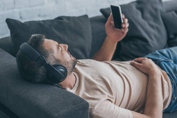 felnőtt ember fejhallgató gazdaság okostelefon üres képernyő, míg pihenő a kanapén - Fotó, kép