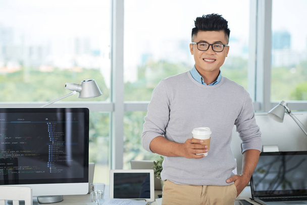 現代のオフィスで様々なデジタル機器で机の近くに立ちながら、熱い飲み物のカップを持ち、笑顔を持つハンサムなアジアの男 - 写真・画像