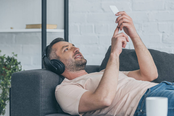 smiling man in headphones lying on sofa and using smartphone - Photo, image