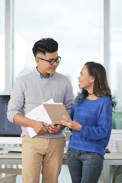 Cheerful Asian man and woman with papers and tablet standing in modern office and discussing design project together - Foto, immagini