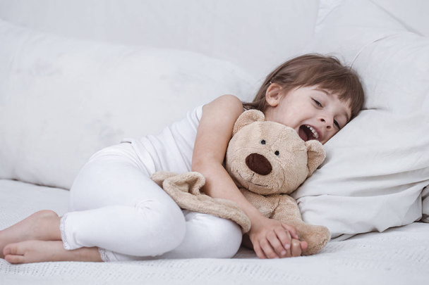 Χαριτωμένο κοριτσάκι χαμογελά ενώ βρίσκεται σε ένα άνετο λευκό κρεβάτι - Φωτογραφία, εικόνα