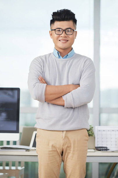 Atrakcyjny azjatycki człowiek uśmiechając się i patrząc na kamerę, stojąc w nowoczesnym biurze i trzymając ramiona skrzyżowane - Zdjęcie, obraz