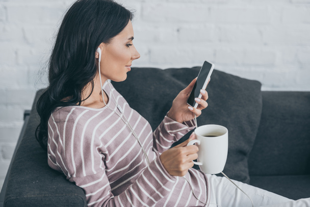 smiling woman using smartphone while listening music in earphones and holding coffee cup - Photo, Image