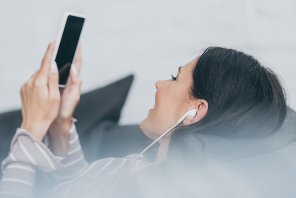 selective focus of young woman listening music in earphones while lying on sofa and holding smartphone with blank screen  - Фото, изображение