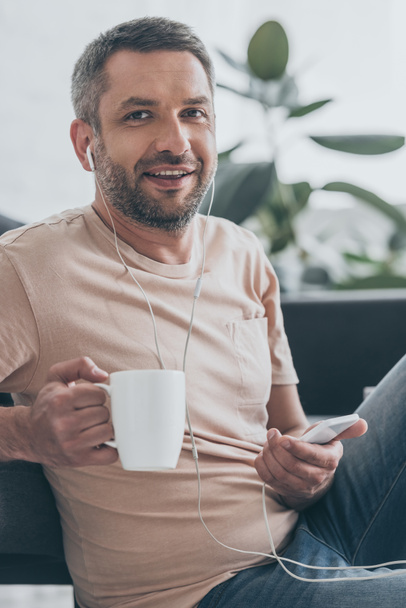 hombre guapo mirando a la cámara mientras escucha música en los auriculares y sosteniendo la taza de café
 - Foto, Imagen