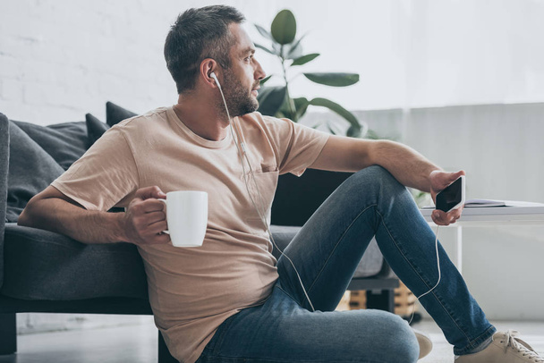 dreamy man looking away while sitting on floor, listening music in earphones and holding coffee cup - Fotó, kép
