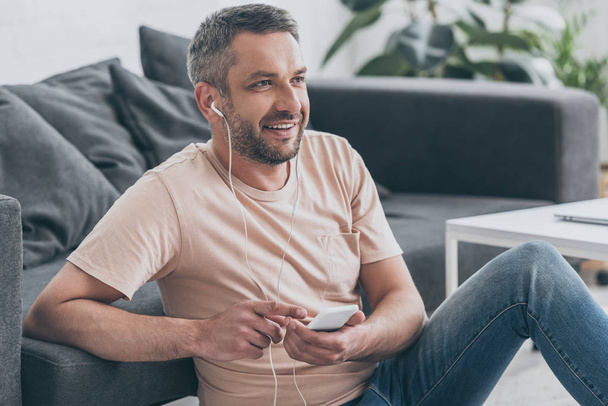 yakışıklı adam kulaklık müzik dinlerken ve evde yerde otururken uzağa bakıyor - Fotoğraf, Görsel