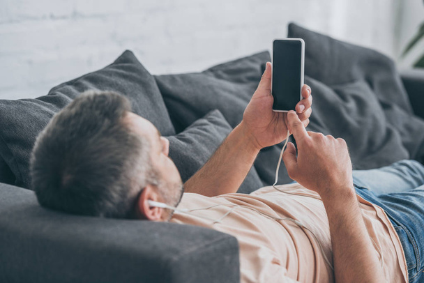 dorosły człowiek słuchania muzyki w słuchawkach i trzymając smartfon z pustym ekranie leżąc na kanapie - Zdjęcie, obraz