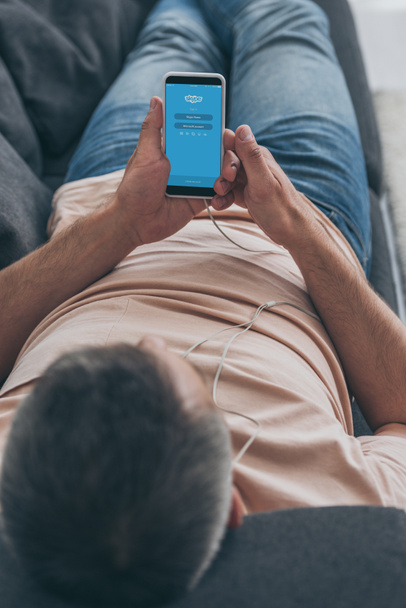 KYIV, UKRAINE - MAY 5, 2019: Selective focus of man in headphones lying on sofa and holding smartphone with Skype app on screen. - Fotoğraf, Görsel