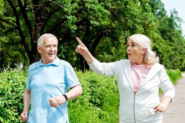 mulher sênior surpreendida apontando com o dedo enquanto corria perto do marido no parque
  - Foto, Imagem