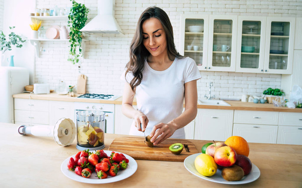 Beautiful woman making fruits smoothies with blender. Healthy eating lifestyle concept portrait of beautiful young woman preparing drink with bananas, strawberry and kiwi at home in kitchen. - Foto, Imagem