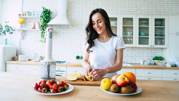 Beautiful woman making fruits smoothies with blender. Healthy eating lifestyle concept portrait of beautiful young woman preparing drink with bananas, strawberry and kiwi at home in kitchen. - Foto, imagen