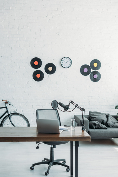 Geräumiges Sendestudio mit Mikrofon und Laptop auf Holztisch, Bürostuhl, Schallplatten und Uhr an weißer Wand - Foto, Bild