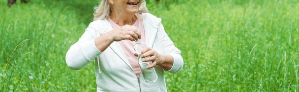 panoramic shot of cheerful senior woman opening bottle with water in park  - Photo, Image