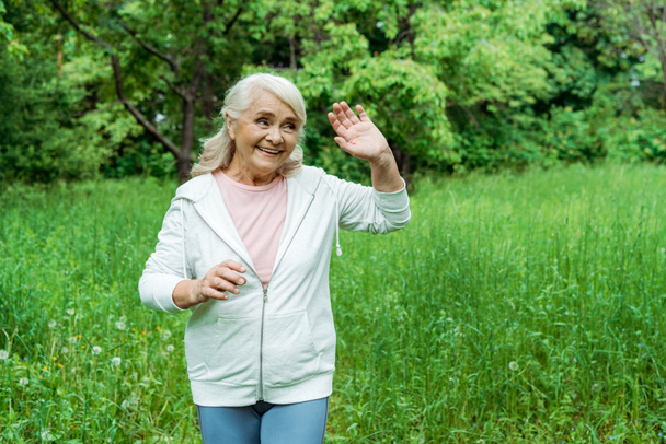 cheerful senior woman with grey hair waving hand in park  - Фото, изображение