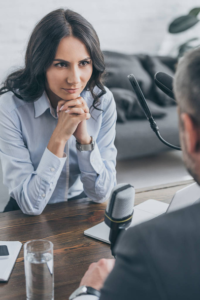 селективный фокус привлекательного радиоведущего, сидящего с сжатыми руками во время интервью с бизнесменом
 - Фото, изображение