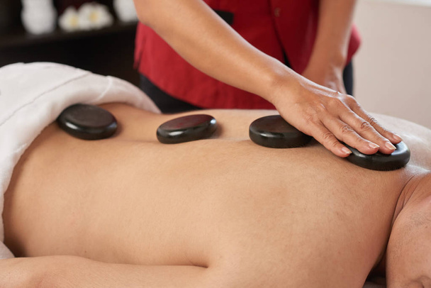 Hands of masseur applying hot basalt stones along spine of man - 写真・画像