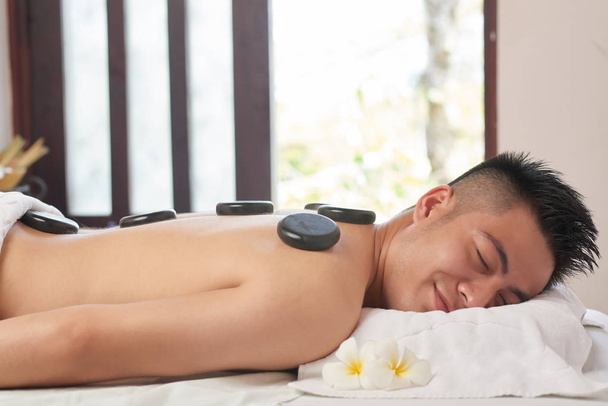Souriant jeune homme asiatique relaxant avec des pierres de massage chaudes sur le dos
 - Photo, image